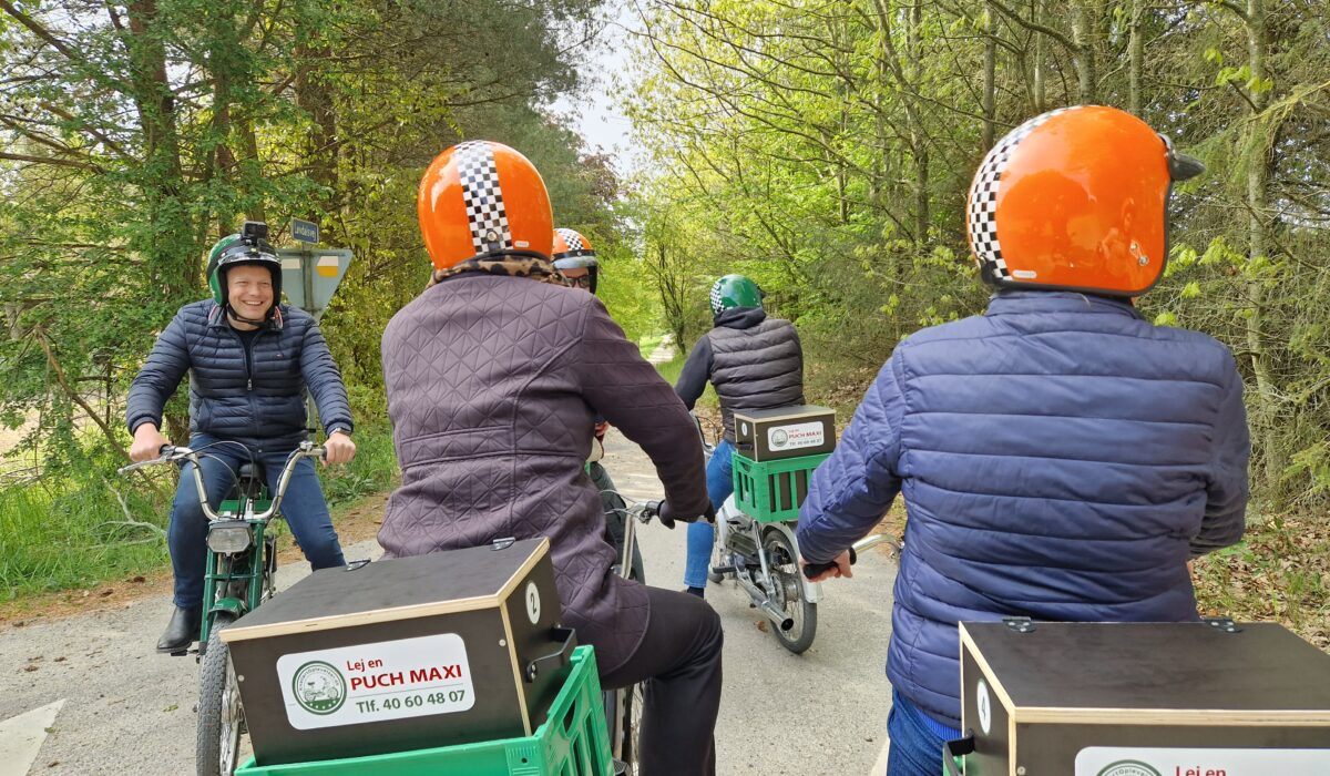 Ryttere med orange ternede hjelme kører på en skovvej, en af dem smiler bagud