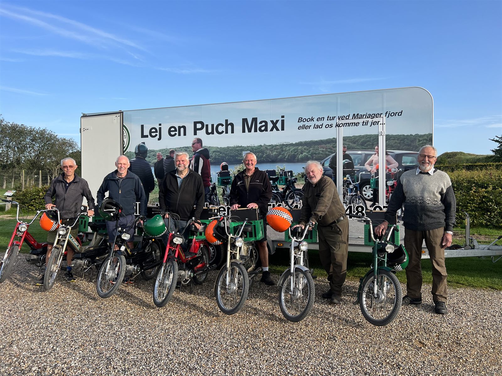Hele flåden af Puch Maxi og en gruppe glade gæster foran traileren med teksten Lej en Puch Maxi